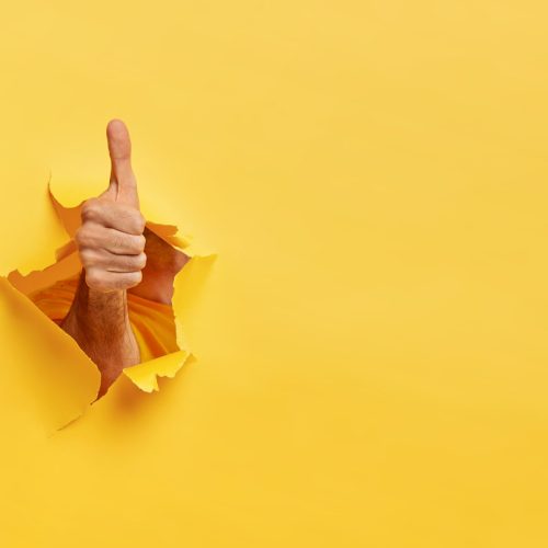 unrecognizable-man-shows-like-gesture-through-torn-yellow-wall-keeps-thumb-up